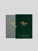 Das Cocktailbuch „Cheers Maus“ neben seinem dunkelgrünen Samtschuber. Die Ansicht verdeutlicht den Kontrast zwischen dem graugrünen Leineneinband des Buches und der weichen Textur des Schubers, beide veredelt mit goldener Titelprägung.