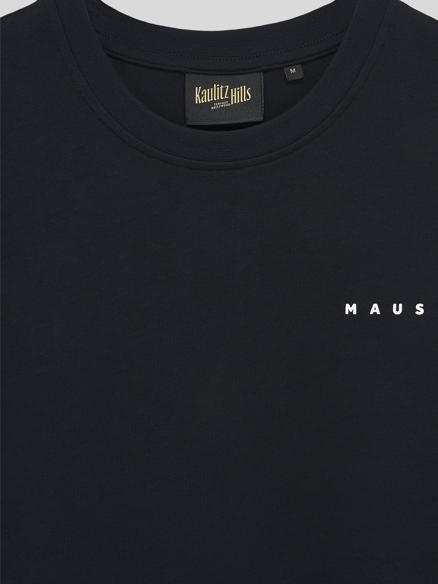 Nahaufnahme der Vorderseite des schwarzen T-Shirts mit kleinem weißen Schriftzug „MAUS“ auf Brusthöhe und Kaulitz Hills Label im Kragen.