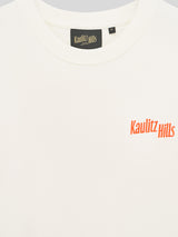 Nahaufnahme des weißen T-Shirts mit kleinem orangefarbenem Stickschriftzug „Kaulitz Hills“ auf der Brust.